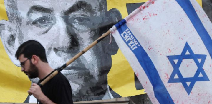 Un manifestante con la bandera israelí pasa junto a un retrato del primer ministro Benjamin Netanyahu durante una protesta antigubernamental que exige la liberación de los rehenes israelíes cautivos desde los ataques del 7 de octubre de 2023 perpetrados por militantes palestinos. Esta protesta se llevó a cabo frente al Ministerio de Defensa en Tel Aviv, en medio de la guerra en curso contra el movimiento militante palestino Hamás.