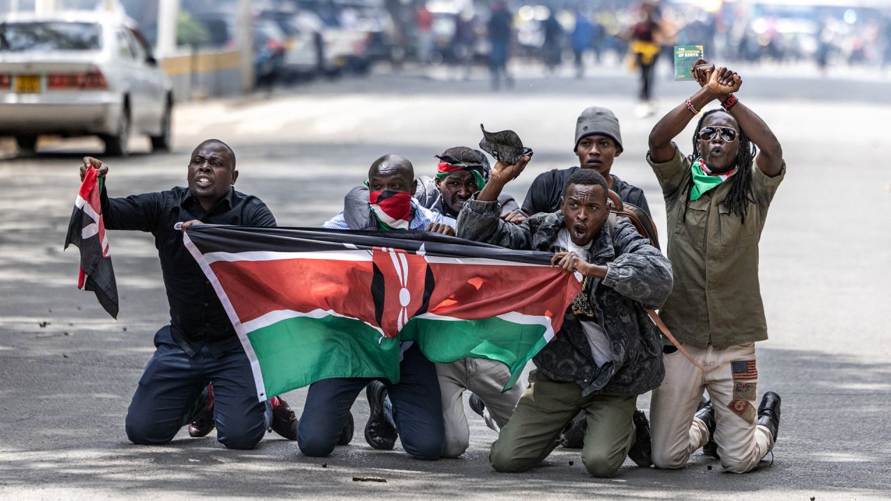 Manifestantes portan una bandera keniana mientras se protegen de los gases lacrimógenos y las balas de goma durante los enfrentamientos entre manifestantes y policías durante una protesta por la muerte del bloguero keniano Albert Ojwang, quien falleció bajo custodia policial, mientras el gobierno presenta el presupuesto 2025-2026 en el centro de Nairobi. | Foto:LUIS TATO / AFP