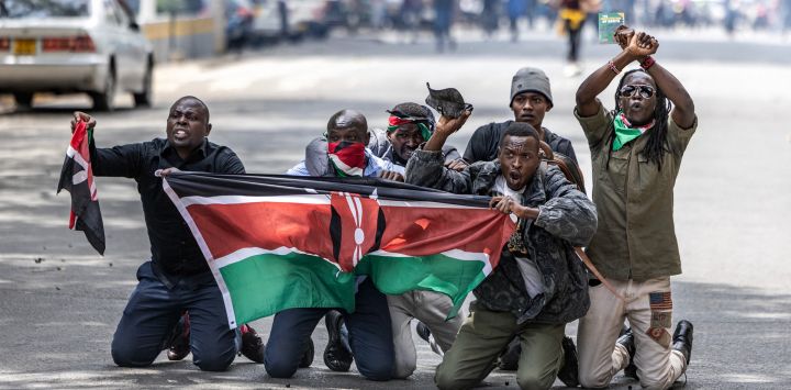 Manifestantes portan una bandera keniana mientras se protegen de los gases lacrimógenos y las balas de goma durante los enfrentamientos entre manifestantes y policías durante una protesta por la muerte del bloguero keniano Albert Ojwang, quien falleció bajo custodia policial, mientras el gobierno presenta el presupuesto 2025-2026 en el centro de Nairobi.