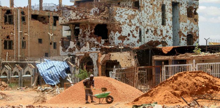 Un trabajador retira piedras de un montículo con una pala y las coloca en una carretilla cerca de unos edificios gravemente dañados en el suburbio de Lamab, en las afueras del suroeste de Jartum, la capital de Sudán, mientras los residentes regresan tras las labores de reconstrucción.