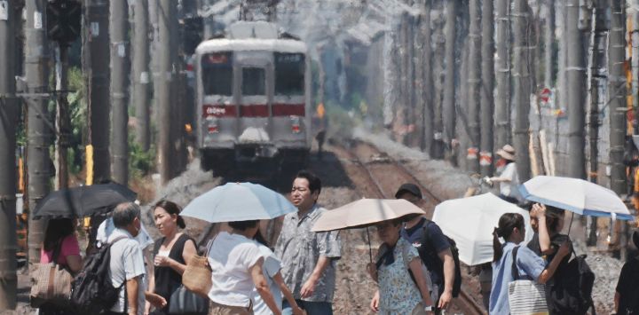 Se ve una neblina de calor al fondo mientras peatones con paraguas cruzan una vía férrea en un día caluroso en Tokio.