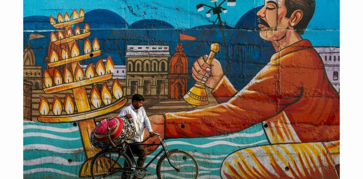 Un hombre lleva un bulto de ropa en su bicicleta junto a un mural en Varanasi, India.