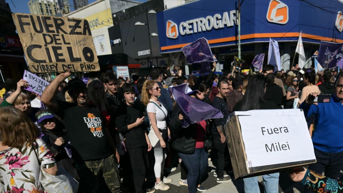An anti-government protester holds a banner against Argentina's President Javier Milei next to supporters of the ruling La Libertad Avanza party as she protests during a motorcade led by Milei in Lomas de Zamora, Buenos Aires Province, on August 27, 2025