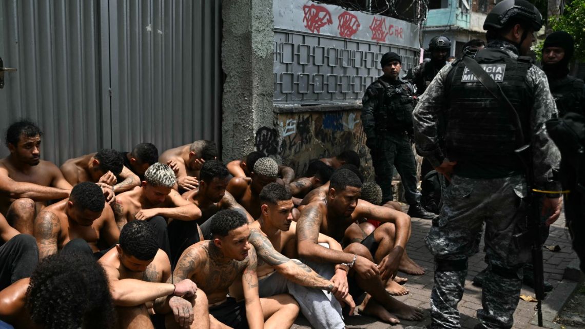 Police officers guard alleged criminals arrested during the Operacao Contencao (Operation Containment) at the Vila Cruzeiro favela, in the Penha complex, in Rio de Janeiro, Brazil, on October 28, 2025. At least 2,500 security forces agents took part in an operation to arrest drug traffickers from the Comando Vermelho (CV), which resulted in 64 people dead, authorities reported. 