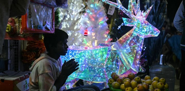 Un vendedor de frutas se sienta junto a un reno navideño iluminado, expuesto a la venta en Bengaluru, India.