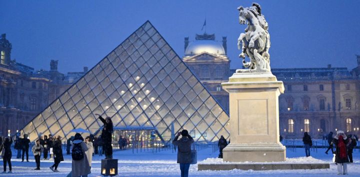Un turista toma una foto de la pirámide del Louvre, diseñada por el arquitecto chino-estadounidense Ieoh Ming Pei, junto a la copia de la estatua "Louis XIV sous les traits de Marcus Curtius" de Bernin, cubierta de nieve en París.