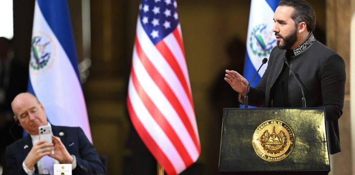 El presidente de El Salvador, Nayib Bukele, pronuncia un discurso ante funcionarios gubernamentales y extranjeros durante el Primer Desayuno Nacional de Oración por El Salvador, mientras el congresista estadounidense Robert Aderholt se toma una foto con su celular en el Palacio Nacional de San Salvador.