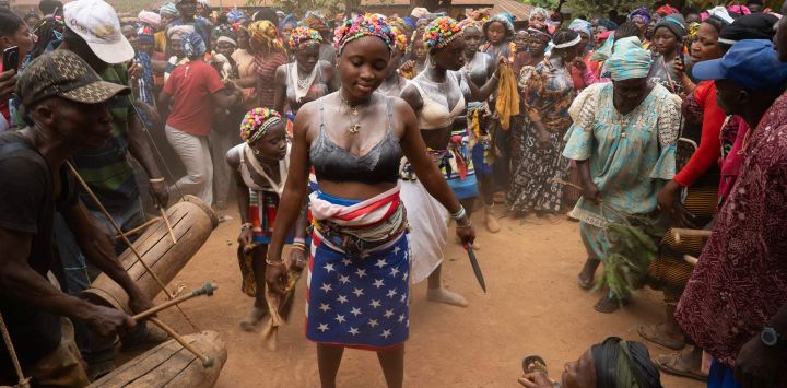 Niñas bailan rodeadas de una multitud en la aldea de Yagala, Sierra Leona. En comunidades de Sierra Leona, la sociedad Bondo organiza un poderoso rito de paso para las adolescentes, uniendo generaciones mediante vibrantes ceremonias que reafirman la identidad cultural. Como se muestra en estas fotografías de la aldea de Yagala, la iniciación comienza con una despedida comunitaria: una celebración dinámica de danza, tambores y bendiciones, donde los ancianos conducen a las niñas hacia el bosque para semanas de aislamiento e instrucción ancestral. Esta práctica, programada para no interrumpir la escuela ni las cosechas, representa una piedra angular de la vida social, marcando su transición a la edad adulta.