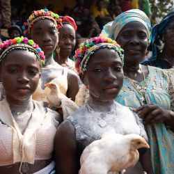 Un grupo de niñas y mujeres líderes de la comunidad y la sociedad Bondo en Sierra Leona posan para una fotografía rodeadas de miembros de la comunidad en la aldea de Yagala. En comunidades de Sierra Leona, la sociedad Bondo organiza un poderoso rito de paso para las adolescentes, uniendo generaciones mediante vibrantes ceremonias que afirman la identidad cultural. Como se muestra en estas fotografías de la aldea de Yagala, la iniciación comienza con una despedida comunitaria: una celebración dinámica con danza, tambores y bendiciones, donde los ancianos conducen a las niñas hacia el bosque para semanas de aislamiento e instrucción ancestral. Esta práctica, programada para no interrumpir la escuela ni las cosechas, representa una piedra angular de la vida social, marcando su transición a la edad adulta. | Foto:Gemma Bonfiglioli / AFP