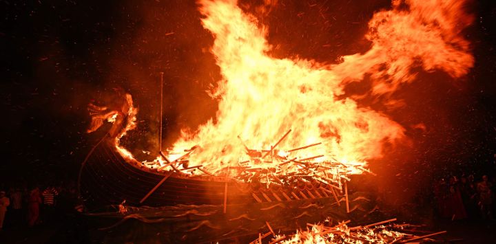 La gente observa el ardor del drakkar vikingo Up Helly Aa "Jarl Squad" en Lerwick, Islas Shetland, durante el festival Up Helly Aa. Up Helly Aa celebra la influencia de los vikingos escandinavos en las Islas Shetland y culmina con hasta 1000 "guizers" (hombres disfrazados) lanzando antorchas encendidas a su drakkar vikingo y prendiéndole fuego al anochecer.