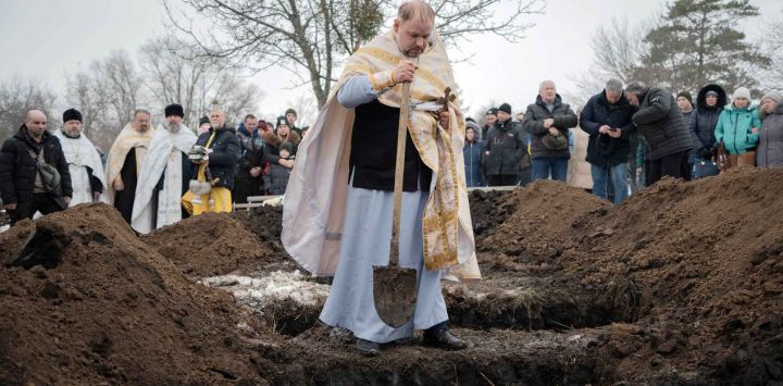 Un sacerdote ucraniano arroja puñados de tierra sobre los ataúdes durante una ceremonia funeraria. Foto de Ivan SAMOILOV / AFP