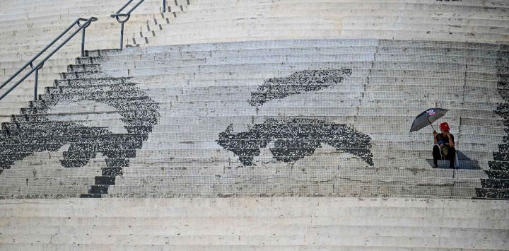 Un hombre cubriéndose del sol con un paraguas descansa en un mosaico que muestra los ojos del fallecido presidente venezolano Hugo Chávez. Foto de Juan BARRETO / AFP