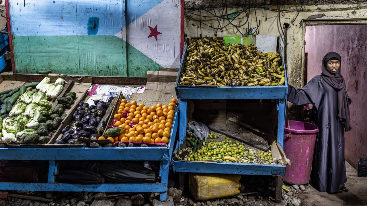Una mujer se encuentra junto a un puesto decorado con la bandera de Yibuti en un mercado al aire libre, mientras los residentes salen a las calles durante las horas más frescas de la tarde para escapar del calor del día en el centro de Yibuti, antes de las elecciones presidenciales de Yibuti de 2026. | Foto:LUIS TATO / AFP