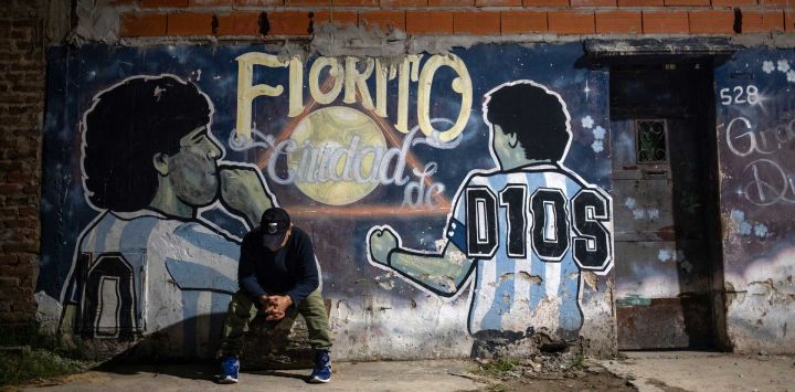 Un hombre se sienta frente a un mural que representa al fallecido astro del fútbol Diego Maradona y lee en español “Fiorito, ciudad de Dios” en el patio de la casa de la infancia del fallecido futbolista, ahora convertida en un comedor social para vecinos necesitados, en Villa Fiorito, provincia de Buenos Aires, Argentina. El segundo juicio por la muerte del astro del fútbol Diego Maradona comienza el 14 de abril de 2026, luego de que el primero, que tuvo lugar el año pasado, fuera anulado al descubrirse que uno de los jueces participaba en un documental clandestino sobre el proceso.
