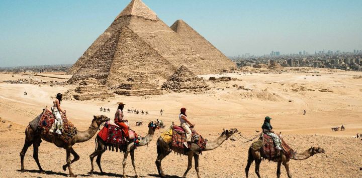 Imagen de turistas paseando en camello en el punto escénico de las Pirámides de Giza, en Giza, Egipto. Las Pirámides de Giza, ubicadas en el suroeste de El Cairo, capital de Egipto, son uno de los sitios más representativos de la antigua civilización egipcia. El complejo incluye las tres pirámides conocidas como las Grandes Pirámides, la imponente escultura conocida como la Gran Esfinge, y varios cementerios antiguos y pirámides más pequeñas.