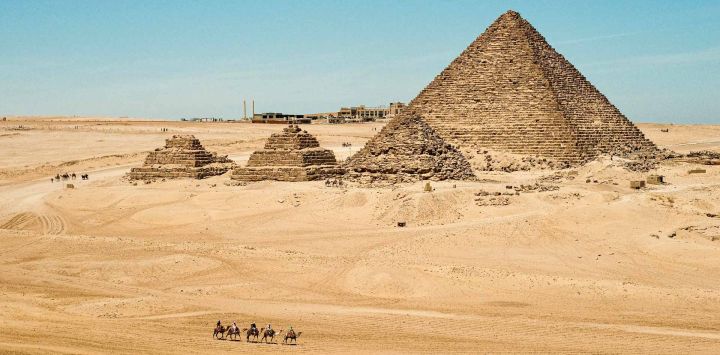 Imagen de turistas paseando en camello en el punto escénico de las Pirámides de Giza, en Giza, Egipto. Las Pirámides de Giza, ubicadas en el suroeste de El Cairo, capital de Egipto, son uno de los sitios más representativos de la antigua civilización egipcia. El complejo incluye las tres pirámides conocidas como las Grandes Pirámides, la imponente escultura conocida como la Gran Esfinge, y varios cementerios antiguos y pirámides más pequeñas.