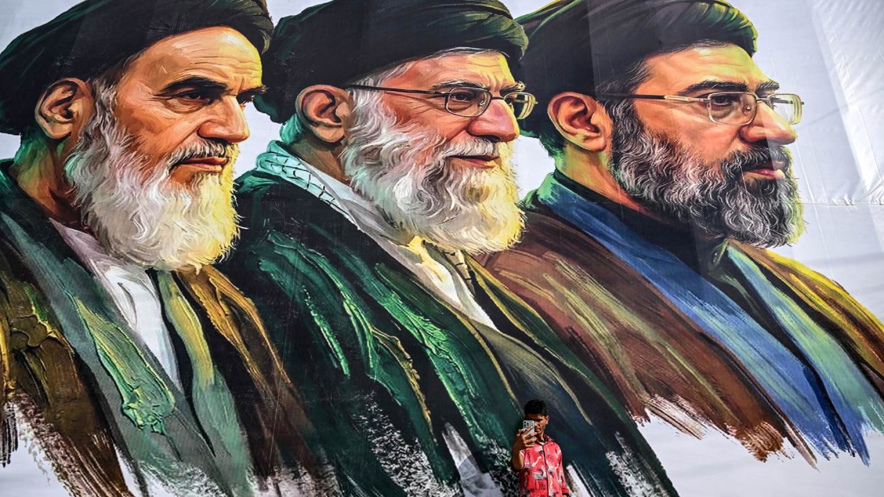 Un hombre se toma una selfie frente a una valla publicitaria que muestra a los difuntos líderes supremos de Irán, el ayatolá Ruhollah Khomeini (izquierda) y el ayatolá Ali Khamenei (centro), junto al recién elegido líder supremo Mojtaba Khamenei, en Srinagar. | Foto:TAUSEEF MUSTAFA / AFP
