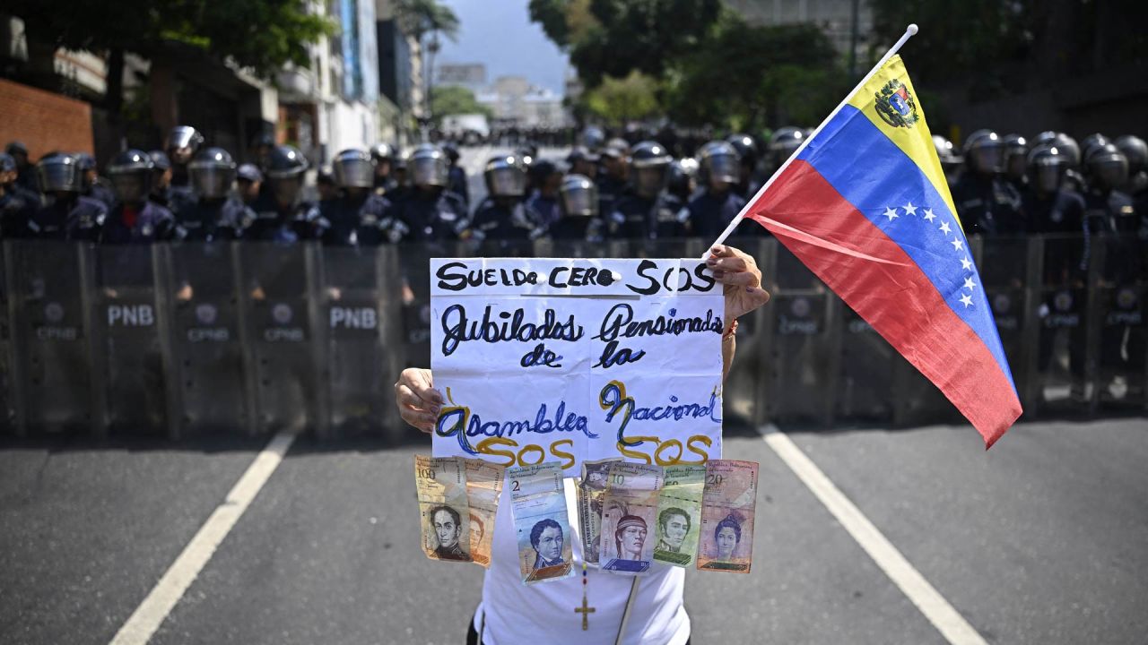 Una mujer sostiene un cartel que dice "Salario cero, SOS jubilados y pensionistas de la Asamblea Nacional" mientras porta una bandera venezolana y billetes de bolívar frente a miembros del Cuerpo Bolivariano de la Policía Nacional (CPNB) durante una manifestación para exigir mejores condiciones laborales y pensiones, en Caracas, Venezuela. | Foto:Federico Parra / AFP