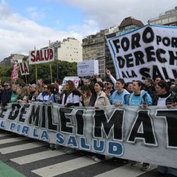 Trabajadores portan una pancarta con el lema "Las medidas de austeridad de Milei son mortales" mientras marchan desde el edificio del Ministerio de Salud (izquierda), cuya fachada luce una imagen de Eva Perón, hasta la Plaza de Mayo durante una protesta en defensa de la salud pública contra los recortes gubernamentales en Buenos Aires. | Foto:JUAN MABROMATA / AFP