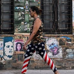Una mujer con leggings con la bandera de Estados Unidos y un perro pasan frente a una casa que exhibe un cartel del fallecido líder venezolano Hugo Chávez, el expresidente cubano Raúl Castro y el fallecido líder revolucionario argentino Ernesto "Che" Guevara, en La Habana, Cuba. | Foto:YAMIL LAGE / AFP