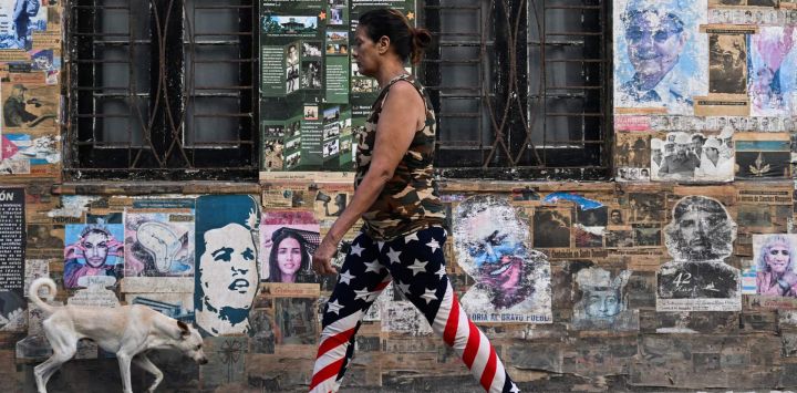 Una mujer con leggings con la bandera de Estados Unidos y un perro pasan frente a una casa que exhibe un cartel del fallecido líder venezolano Hugo Chávez, el expresidente cubano Raúl Castro y el fallecido líder revolucionario argentino Ernesto "Che" Guevara, en La Habana, Cuba.