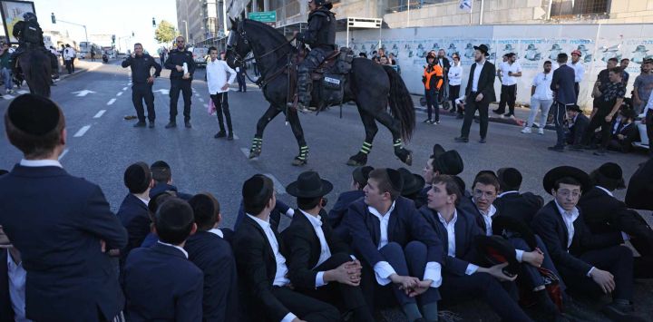 Policías israelíes montan guardia mientras judíos ultraortodoxos protestan contra el servicio militar obligatorio bloqueando la entrada a Jerusalén. Se oponen a una modificación de la ley sobre el servicio militar obligatorio, del que la comunidad ultraortodoxa ha estado tradicionalmente exenta. Según una disposición establecida cuando se fundó el país en 1948 —cuando la comunidad ultraortodoxa era pequeña—, los hombres que se dedican a tiempo completo al estudio de los textos sagrados judíos están exentos.