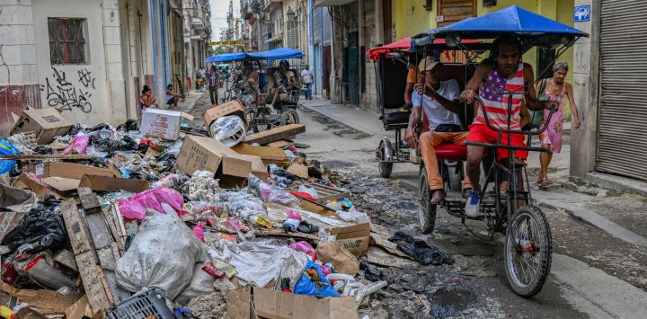 Una persona con una camiseta con la bandera de Estados Unidos pasa en triciclo junto a un montón de basura en una calle de La Habana, Cuba.