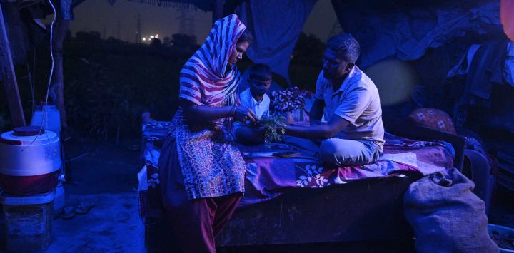 Esta fotografía muestra a Debvati (izquierda) conversando con su esposo, el agricultor Bhole Shankar, dentro de su humilde vivienda al anochecer, en las llanuras aluviales del río Yamuna, en Nueva Delhi. En las exuberantes riberas verdes de las llanuras aluviales del río Yamuna, en Nueva Delhi, los veranos abrasadores se han vuelto cada vez más difíciles de soportar, lo que exige adaptaciones y obliga a la vida a transcurrir durante las horas de oscuridad antes de que el sol se vuelva implacable. La capital de la India alcanzó los 46,5 °C este verano, todavía por debajo del récord de 49,9 °C registrado en 2024.