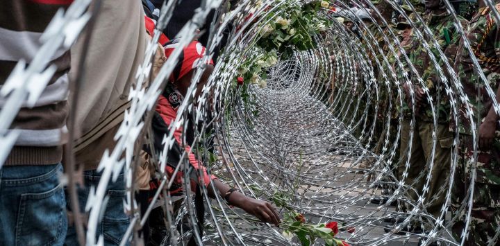 Agentes de la policía keniana observan cómo activistas colocan flores en la barrera de alambre de púas instalada para impedirles el acceso al recinto del Parlamento durante las manifestaciones que conmemoran el segundo aniversario de las mortales protestas juveniles contra la Ley de Finanzas de junio de 2024, en las que los manifestantes irrumpieron en el Parlamento, en Nairobi.