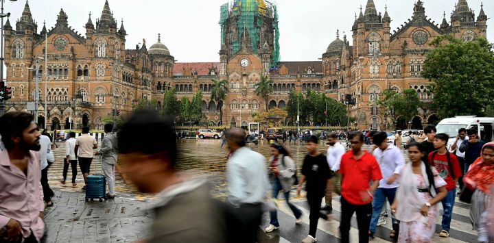 Esta fotografía muestra a peatones cruzando una calle para acceder a una acera cerca de la estación de tren Chhatrapati Shivaji Maharaj Terminus (CSMT) en Bombay. En India, una acera puede tener muchos usos —como un puesto ambulante, un aparcamiento o un vertedero—, pero rara vez cumple su función original: ser un camino seguro y despejado para caminar.