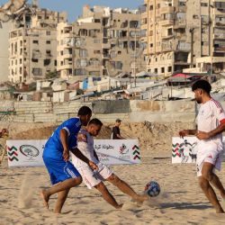 Palestinos desplazados del equipo Shati (azul) y del Club Deportivo de Gaza (blanco) participan en un torneo de fútbol playa en la costa de la ciudad de Gaza, con edificios destruidos y dañados al fondo. | Foto:OMAR AL-QATTAA / AFP