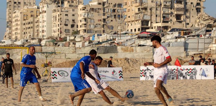 Palestinos desplazados del equipo Shati (azul) y del Club Deportivo de Gaza (blanco) participan en un torneo de fútbol playa en la costa de la ciudad de Gaza, con edificios destruidos y dañados al fondo.