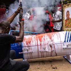 Un hombre dispara un arma de fuego tradicional junto al ataúd ensangrentado con forma de pluma del difunto maestro ghanés Samuel Commey Tetteh, después de que se sacrificara una oveja sobre él como parte de los rituales tradicionales durante su funeral en Paanor, Ghana. Los ataúdes de fantasía son una tradición funeraria ghanesa distintiva, particularmente asociada con el pueblo Ga de la región del Gran Accra. Conocidos localmente como abebuu adekai ("cajas de proverbios"), se elaboran a mano con la forma de objetos, animales o vehículos que reflejan la profesión, la personalidad o los intereses del difunto, desde un pez para un pescador hasta un zapato para un futbolista. | Foto:OLYMPIA DE MAISMONT / AFP