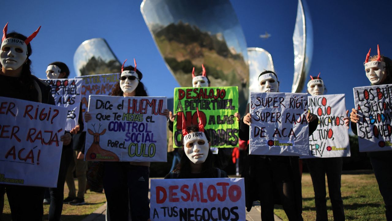 Manifestantes con máscaras y pancartas participan en una protesta contra Peter Thiel, presidente de Palantir Technologies, junto al monumento Floralis Generica en Buenos Aires. | Foto:Emiliano Lasalvia / AFP