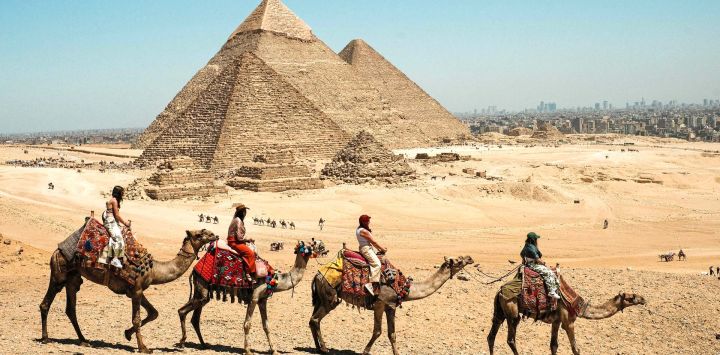 Imagen de turistas montando camellos en el complejo de las pirámides de Giza, en Egipto. Egipto es la cuna de una antigua civilización que floreció a lo largo del río Nilo. Desde las pirámides que han permanecido en pie durante milenios hasta sus bulliciosas ciudades modernas, lo antiguo y lo contemporáneo se entrelazan por todo el país, reflejando una civilización que ha perdurado a través de los siglos.