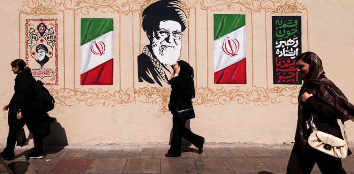 Unos iraníes pasan frente a un muro decorado con la imagen del asesinado líder supremo de Irán, el ayatolá Alí Jamenei, y la bandera nacional, en el centro de Teherán. Estados Unidos e Israel lanzaron ataques contra Irán el 28 de febrero, matando al líder supremo iraní y a altos mandos militares, lo que llevó a las autoridades a tomar represalias con ataques contra Israel y en toda la región del Golfo.