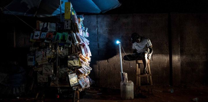 Un vendedor utiliza una batería portátil para iluminar su puesto durante un corte de electricidad en Westfield, en medio de apagones prolongados que han alterado la vida cotidiana en todo el país. Desde junio, esta pequeña nación de África Occidental se ha visto gravemente afectada por apagones prolongados, cuya intensidad ha alcanzado niveles críticos en las últimas semanas, justo en el punto álgido de la temporada de calor. Con una duración que a veces oscila entre las 12 y las 48 horas, estos cortes han sumido a los gambianos en una oscuridad sofocante durante la noche y han supuesto la pérdida del sustento para amplios sectores de la sociedad. La semana pasada estallaron dos noches de protestas caóticas y espontáneas, en las que los manifestantes exigían la destitución del presidente.