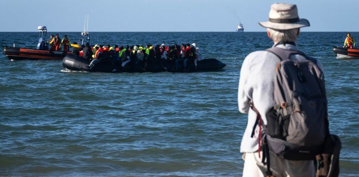Un turista observa a migrantes a bordo de una embarcación de traficantes que intentan cruzar el canal de la Mancha bajo la vigilancia de los servicios de rescate franceses, frente a la playa de Merville-Franceville-Plage, en el noroeste de Francia. A medida que las travesías por el canal de la Mancha se vuelven cada vez más difíciles desde las playas del norte de Francia, los migrantes prueban nuevas rutas para llegar a Inglaterra desde regiones del sur, como el departamento de Calvados, donde el canal es más ancho que a lo largo de la costa septentrional francesa.