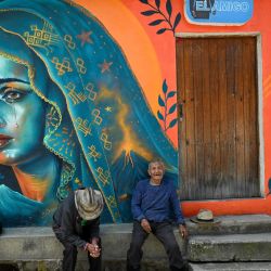 Dos hombres indígenas descansan frente a un mural de la Virgen de los Dolores en las calles de Santa María de Jesús. Los murales forman parte de "Un nuevo aire para Santa María de Jesús", una iniciativa que busca restaurar espacios deteriorados y fortalecer la identidad de esta comunidad a través del arte. | Foto:Johan Ordóñez / AFP
