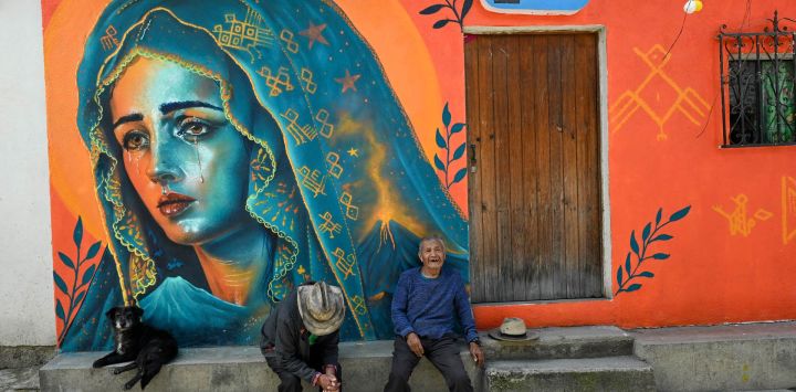 Dos hombres indígenas descansan frente a un mural de la Virgen de los Dolores en las calles de Santa María de Jesús. Los murales forman parte de "Un nuevo aire para Santa María de Jesús", una iniciativa que busca restaurar espacios deteriorados y fortalecer la identidad de esta comunidad a través del arte.