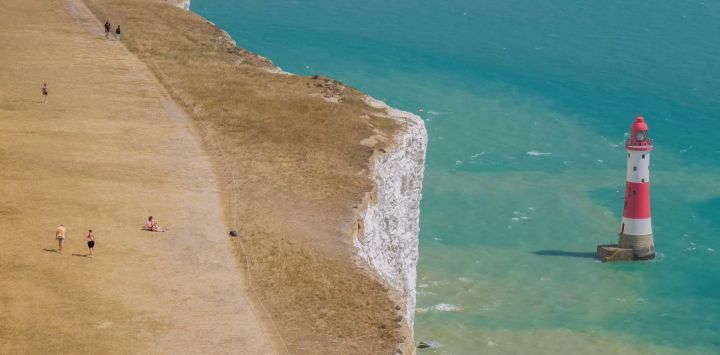 Esta vista aérea muestra a personas caminando sobre hierba completamente seca, a lo largo del borde del acantilado sobre el faro de Beachy Head, cerca de Eastbourne, en la costa sur de Inglaterra.