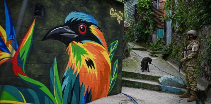 Un soldado salvadoreño monta guardia en un pasaje estrecho de la comunidad 10 de Octubre, frente a un mural pintado por el artista Carlos Clímaco, en San Marcos, El Salvador. El proyecto artístico busca transformar la identidad de una comunidad históricamente afectada por la violencia de las pandillas.